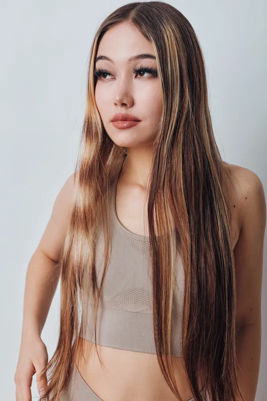 Young woman with long straight hair and neutral makeup wearing beige crop top standing against light background with serious expression looking aside.