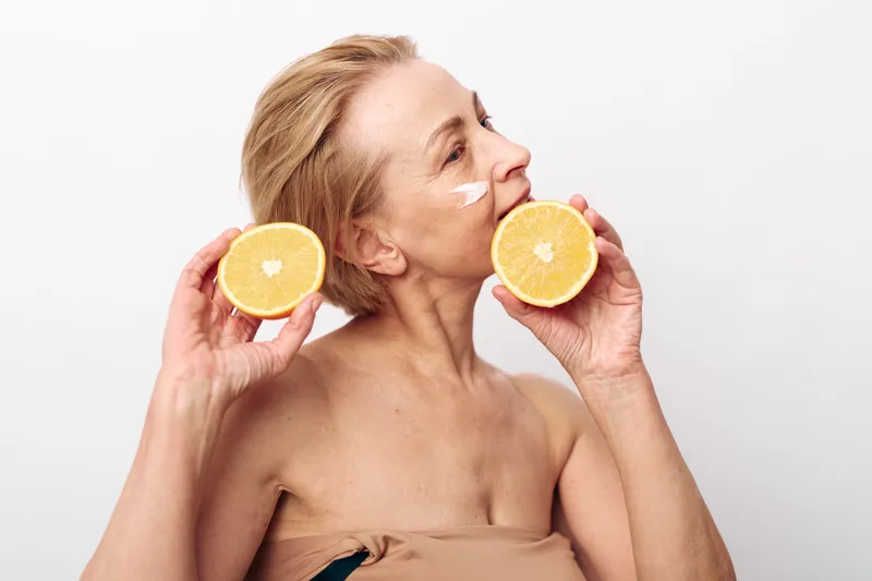 Mature woman with natural skin holds two halves of a citrus fruit near her face, applying facial cream on her cheek in a bright studio. Concept of skincare, natural beauty, and healthy lifestyle.