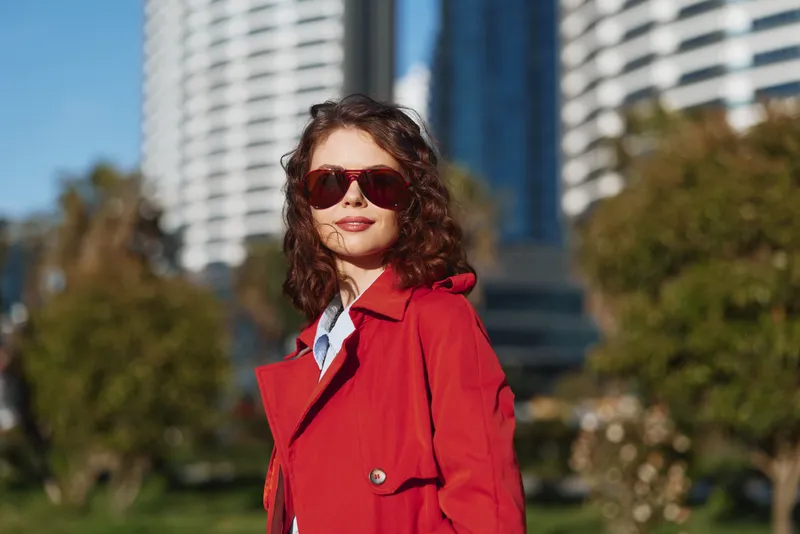 A young woman in a red trench coat stands in an urban park, wearing sunglasses, posing confidently as modern skyscrapers loom behind.