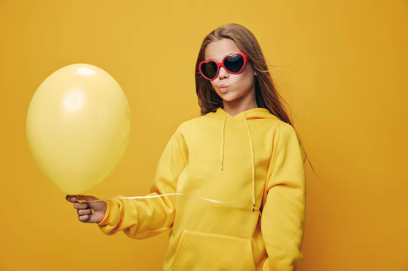 Girl in yellow hoodie and red heart shaped sunglasses holding yellow balloon with pouting lips on yellow background for party fun and celebration mood.