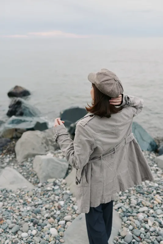 Woman in grey coat and hat stands on rocky shore looking at calm sea, back view. Casual autumn outfit, peaceful coastal scene, concept of solitude and nature relaxation.