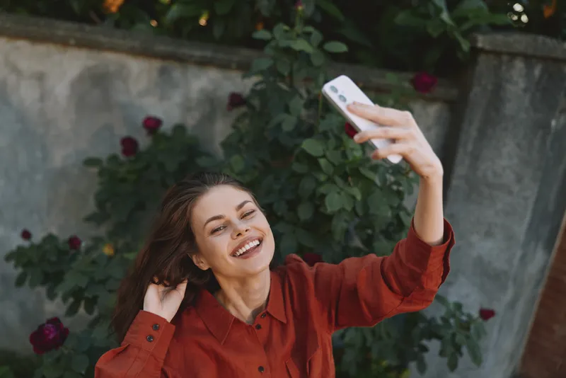Cheerful woman outdoors, taking a selfie with warm sunlight, wearing a red shirt, smiling with relaxed posture, lush greenery behind, vibrant natural colors and carefree lifestyle