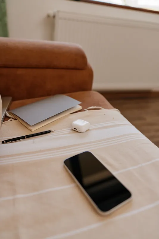 A tidy home table setup with a smartphone, notebook, pen and wireless earbuds on a light checkered tablecloth in a cozy living room, conveying calm daily routine