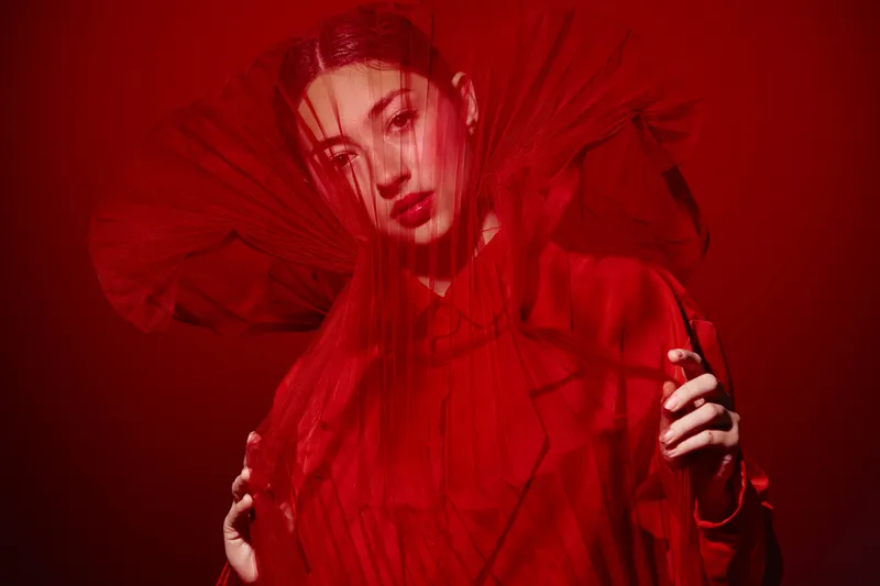 Portrait of a woman in a detailed red fashion dress and veil posing against a red background. Creative beauty concept with dramatic mood and fabric texture.