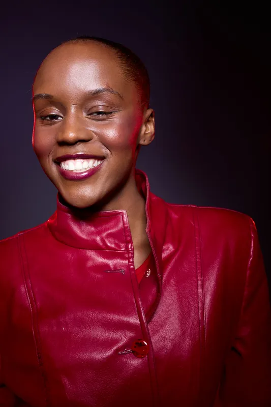 Smiling African woman with dark skin wears red leather jacket, posing confidently against dark background. Concept for fashion, beauty, confidence, and empowerment.
