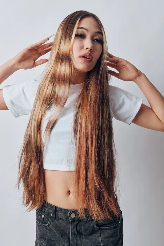 Young woman with long straight hair wearing white crop top and black pants posing on a bright background with hands near head for hair styling and fashion concept.