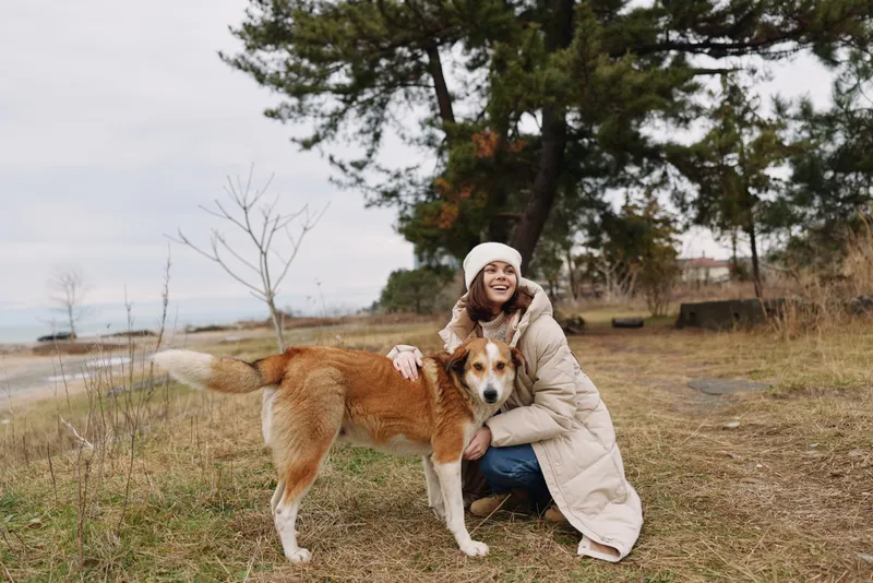 Woman kneels in a warm coat at a park, embracing a friendly dog on grass with trees in the background, capturing wholesome companionship and a serene outdoor moment.