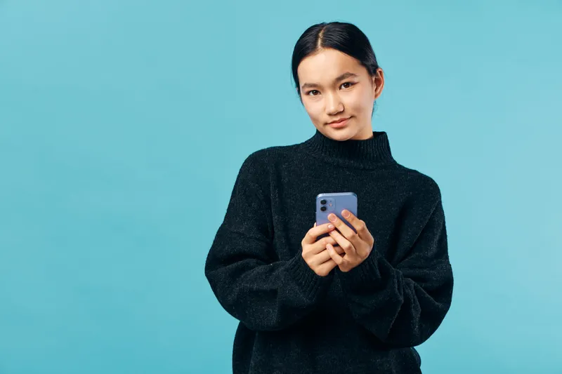Young woman holding smartphone wearing black turtleneck sweater standing against blue background looking at camera with slight smile in portrait indoors studio lighting casual style.