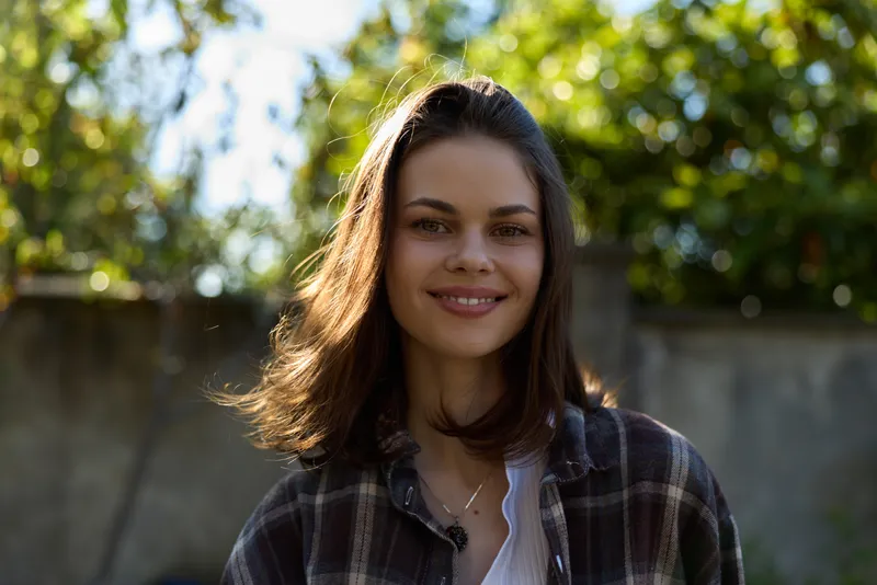 Smiling woman with shoulder length hair wearing a plaid shirt outdoors. Close-up portrait against a natural green background. Concept of lifestyle, wellness, and casual style