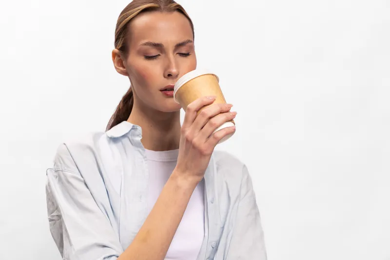Young woman with closed eyes smells hot beverage in disposable cup isolated on white background. Concept of coffee aroma, relaxation, and morning routine for healthy lifestyle.