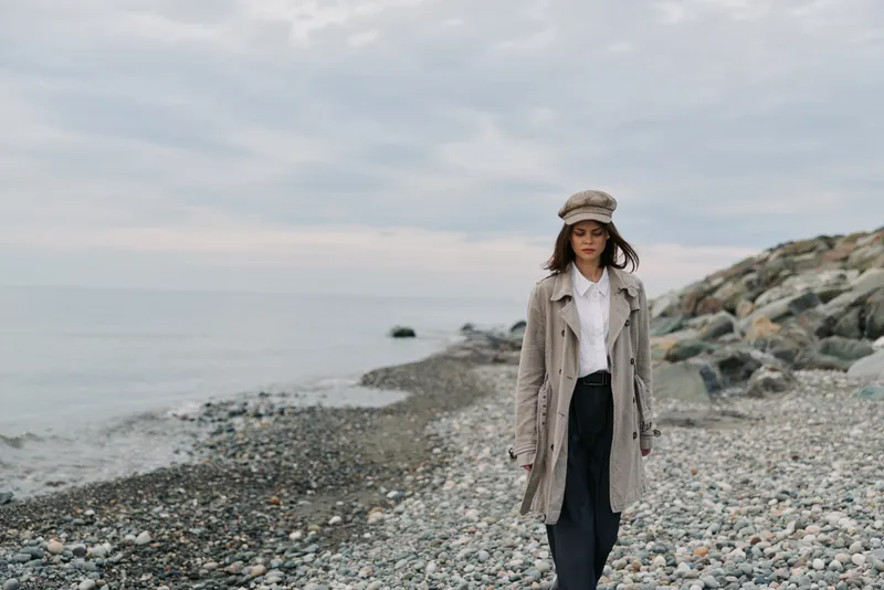 Woman in beige coat and hat walks on rocky beach under cloudy sky. Calm sea and stones create peaceful atmosphere. Concept of autumn fashion and outdoor lifestyle.