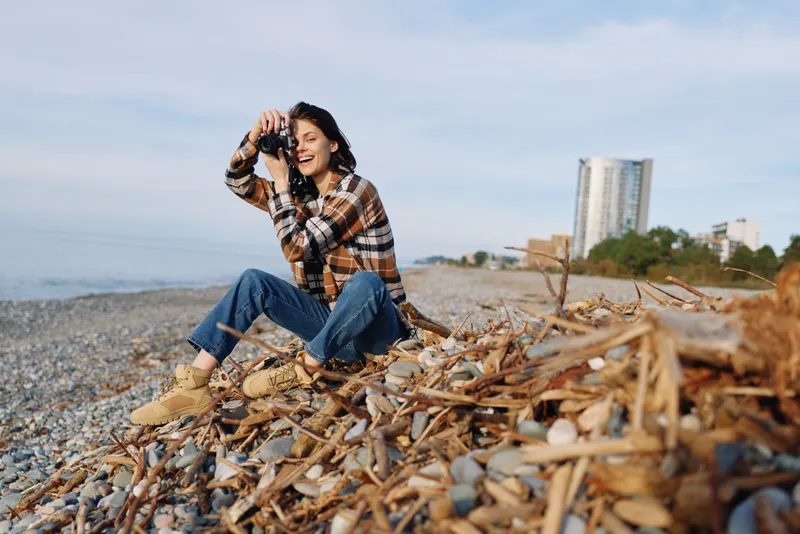 Woman outdoors on a beach, sitting among driftwood, using binoculars, casual plaid shirt and jeans, calm sky and distant cityscape, natural coast scene, travel mood, candid nature moment