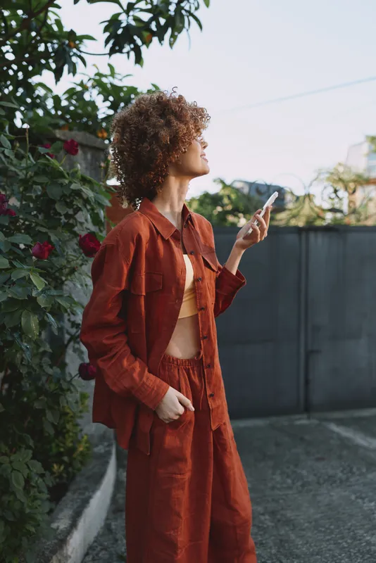 Young woman with curly hair in a rust jumpsuit stands outdoors among roses, looking up and reaching toward the air in a casual fashion moment