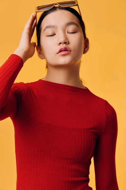 Young woman in red sweater poses with closed eyes and hand on head against yellow background for fashion portrait and beauty concept in studio photo session.