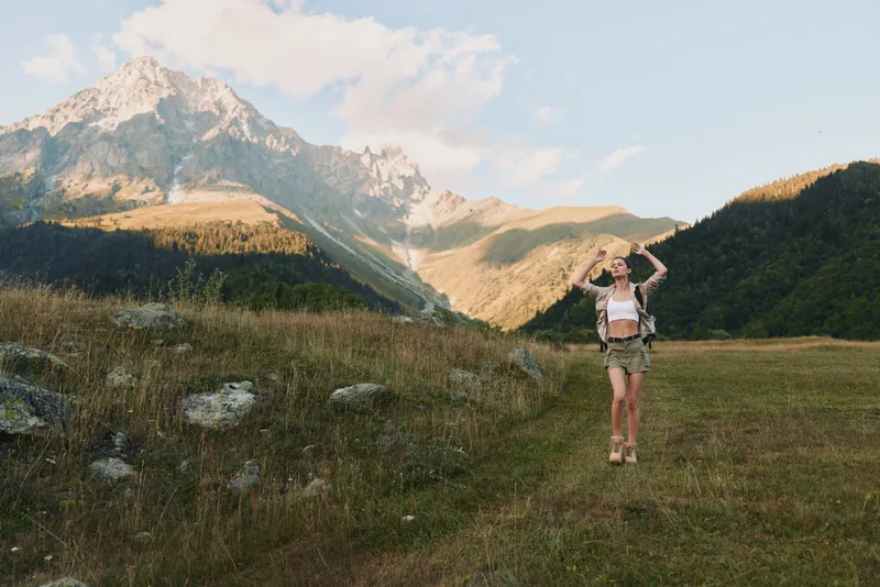 Cheerful woman outdoors, hiking in alpine meadow, sunlit moment, relaxed posture, light breeze, natural colors, mountain scenery, travel vibe, nature immersion