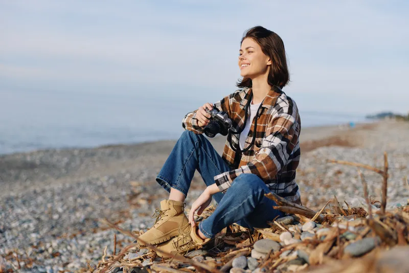 Outdoor woman sits on a pebbled beach with boots and a plaid shirt, smiling at the sea breeze, enjoying calm coastal scenery, a relaxed moment of nature and photography by the shore