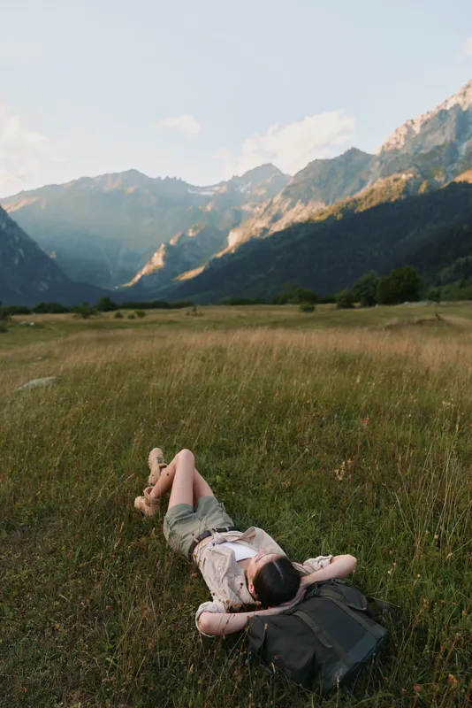 Young woman relaxing in a sunlit meadow with distant mountains, lying on grass with a backpack, capturing a peaceful, carefree moment