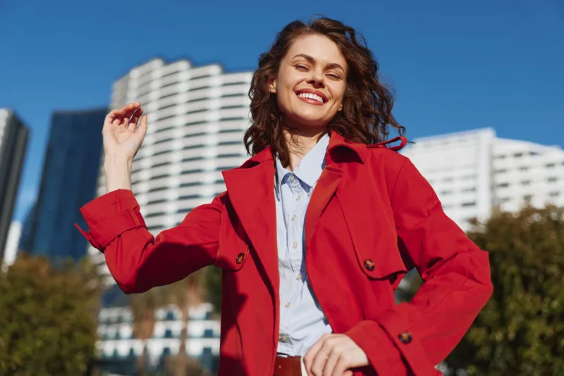 Young woman in a red coat smiles in a sunny city square, projecting confidence and style as tall buildings rise behind her, creating a bold urban mood with warm light