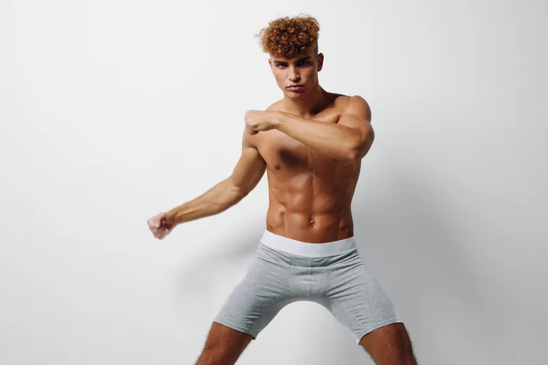 Young muscular man with curly hair posing shirtless in gray shorts on white background, showing fitness, strength, and athletic body in studio lighting for health and sport concept.