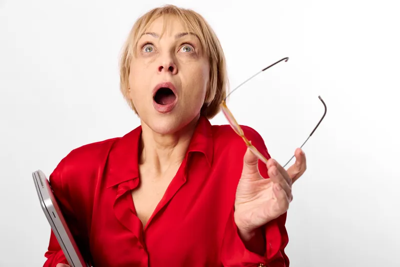 Woman surprised in red shirt holding glasses, shocked expression, open mouth and wide eyes, studio portrait on white background, close up, clean copyspace