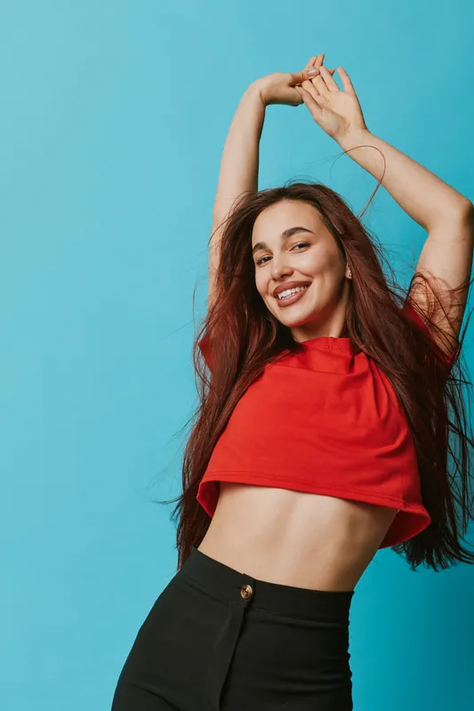 Young woman with long hair wearing red crop top and black pants posing with raised arms and smiling on blue background. Casual fashion and confident model with natural look.