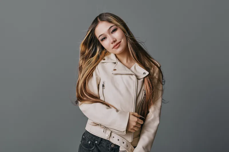 Young woman with long hair wearing beige leather jacket posing with closed eyes against gray background in studio for fashion portrait and beauty concept.