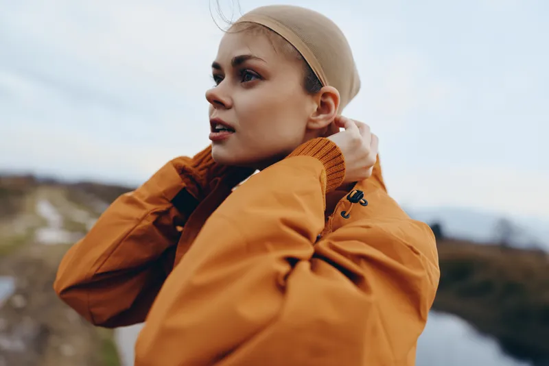 Woman lifestyle outdoors jacket hat wind profile nature travel mountains in soft daylight A young woman in an orange jacket and beige hat turns in profile with hair lifted by wind against a distant