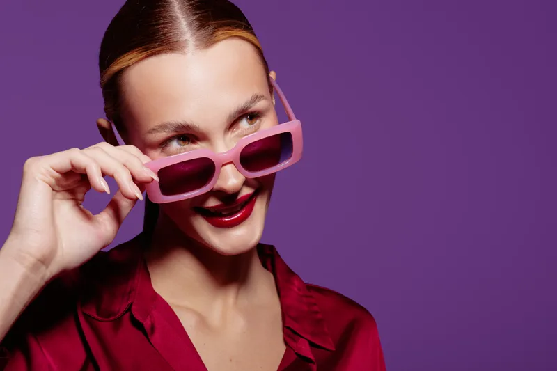 Young woman in red blouse wearing pink sunglasses looks sideways with a smile on purple background. Concept of fashion, style, confidence, and modern accessories.