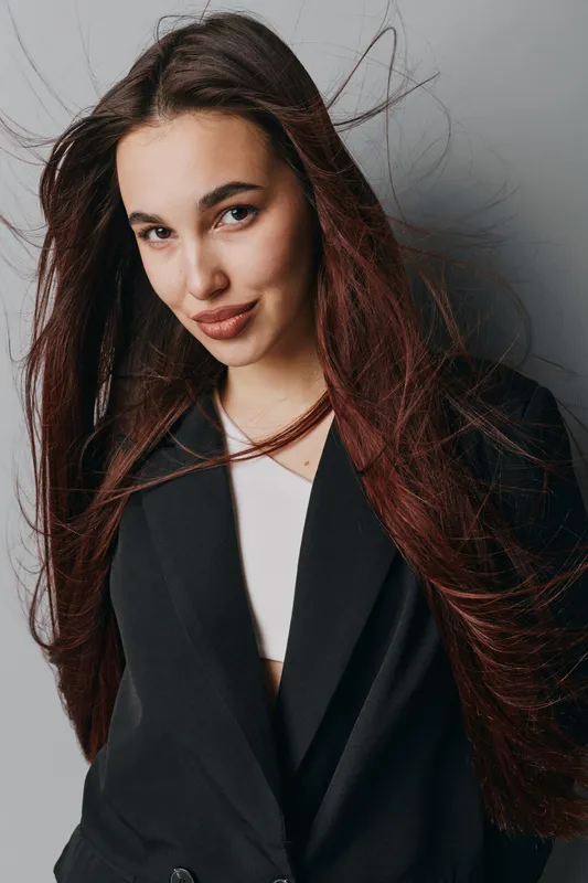 Young woman with long hair wearing black blazer and white top posing against gray background. Portrait of confident female with natural makeup and relaxed expression in studio lighting.