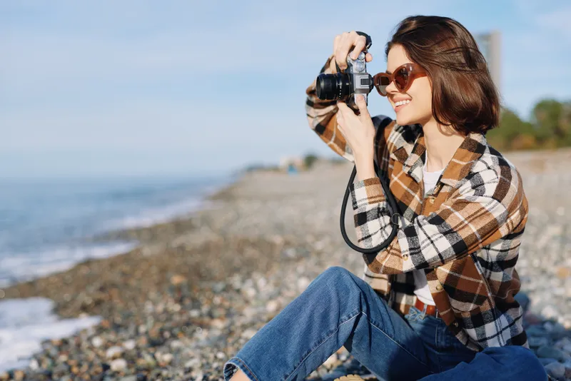 Photographer woman on a pebble beach takes a photo with a camera, wearing a plaid shirt, jeans and sunglasses, enjoying outdoor shooting by the sea today