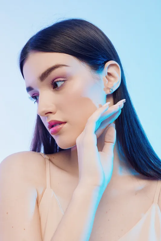 Young woman with long dark hair poses with hand near face in soft studio lighting. Beauty and fashion concept with delicate makeup and smooth skin on blue background.