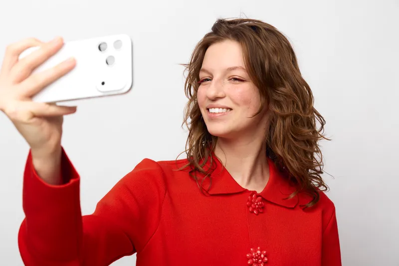 Young woman with curly hair wearing red sweater takes selfie with smartphone on white background. Concept of casual lifestyle, technology, and self-portrait photography.