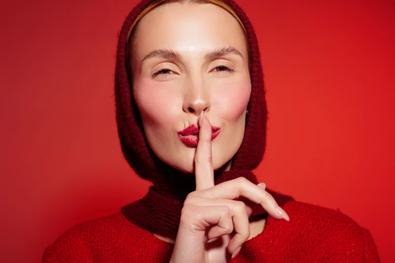 Woman in red sweater and hood makes silence gesture with finger on lips against red background. Concept of secrecy, quiet, and calm expression in studio portrait.