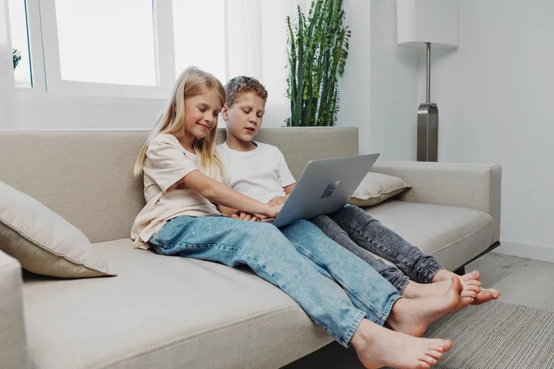 Two children sitting barefoot on a couch using a laptop together in a bright living room with casual clothing and relaxed mood.