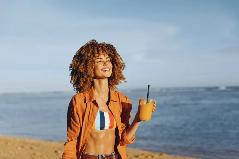 Cheerful woman outdoors by the seaside, curly hair, orange jacket, holding glass with orange juice, sunny weather, smiling, relaxed posture, natural light, calm ocean background