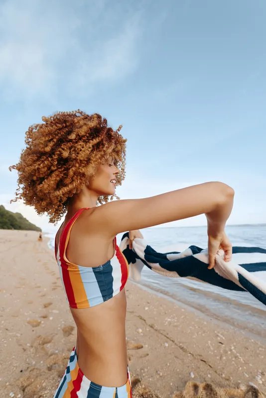 Young woman with curly hair adjusts a striped swimsuit on a sandy beach as she holds a towel, the ocean and a clear blue sky framing the scene, conveying a playful summer vibe.