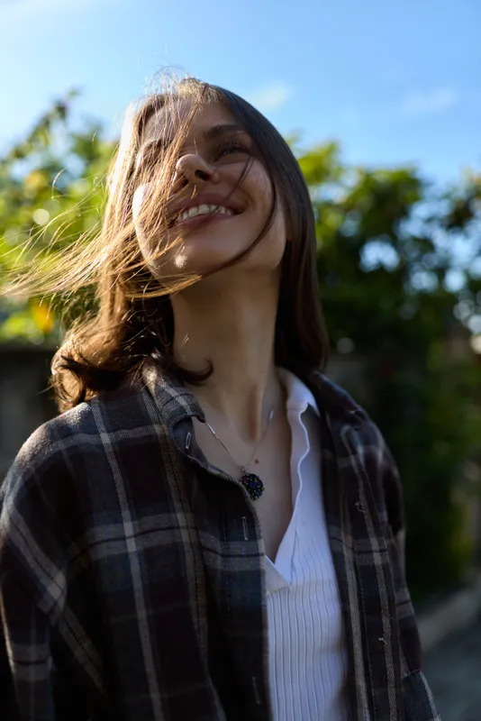 Smiling woman outdoors, lifestyle focused, plaid shirt, sunlight, casual pose, portrait in nature, conveying warmth, wellness, confidence, and everyday happiness with soft natural light