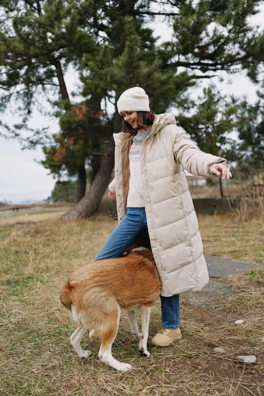 Woman in a long beige winter coat and beanie walks with a large dog in a park, grass and pine trees in the background, candid outdoor moment of companionship and exploration