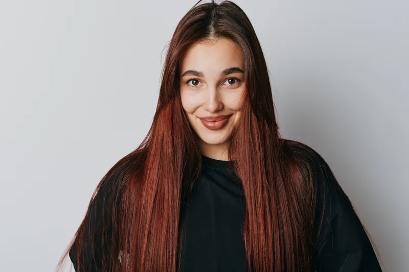 Young woman with long straight hair smiling and looking at camera on white background. Casual style, natural makeup and confident expression for portrait photography.