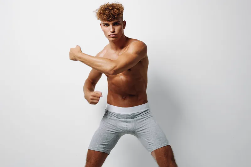 Young athletic man with curly hair posing shirtless in gray shorts against white background for fitness, strength, and active lifestyle concept in studio lighting.