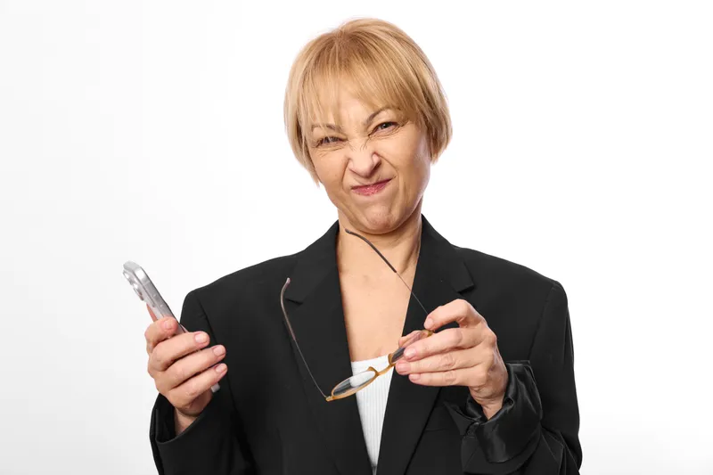 Elderly woman smiling with short blond hair holding smartphone and glasses, isolated on white background, portrait of communication, everyday technology, friendly mood