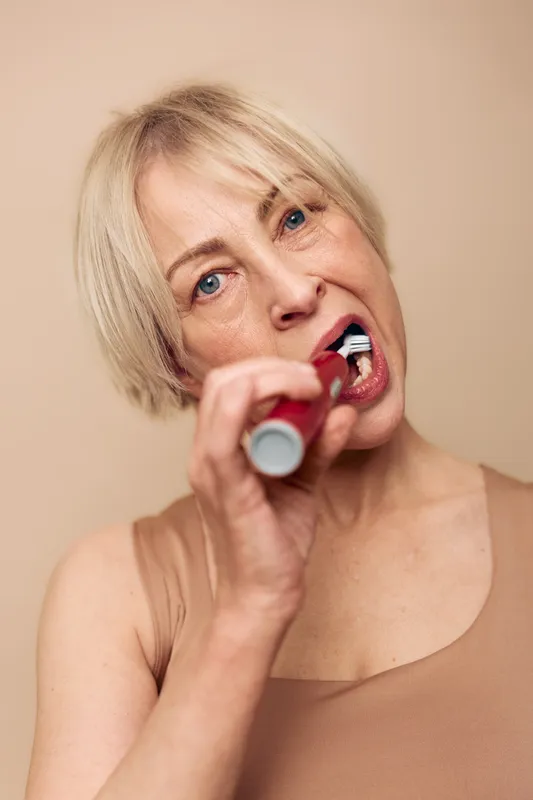 Middle aged woman with short blonde hair cleans teeth with electric toothbrush against beige background. Concept of dental hygiene, oral care, healthy habits, and personal wellness routine.