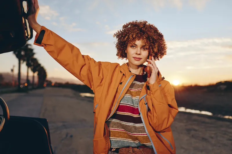 Woman with curly hair wearing an orange jacket on a city street at sunset, standing by a parked car with one arm raised, lifestyle mood, warm golden light