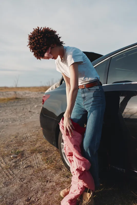 Car, person, outdoor, denim, jacket, standing beside a car in a dusty landscape, casual fashion moment with wind moving fabric and sunlit plains for a candid street style shot