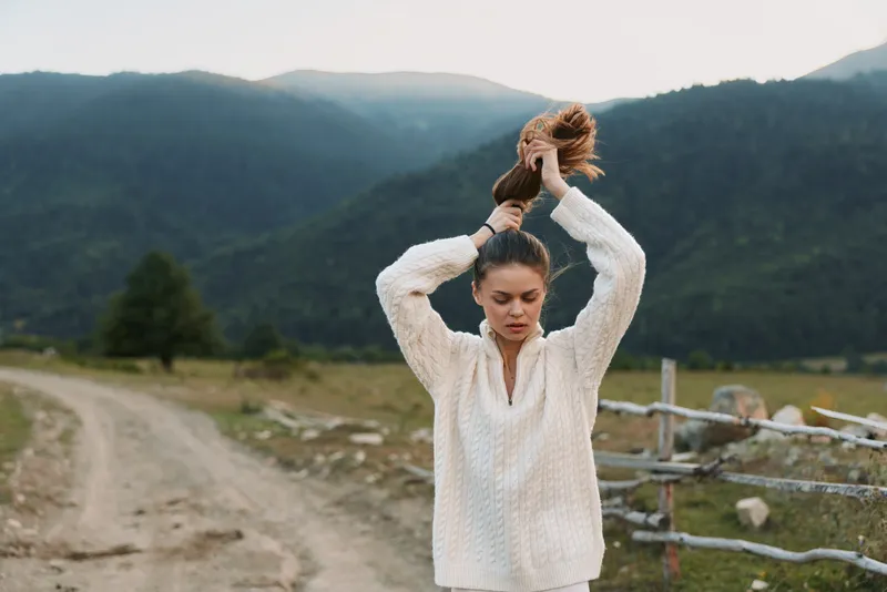 Cheerful woman outdoors, hair tied back, wearing a cozy sweater, relaxed pose in a countryside setting, soft breeze, natural light, calm atmosphere amid wide fields