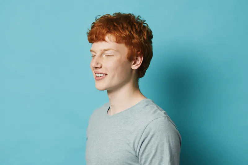 Young man with red hair and freckles wearing gray t shirt standing with eyes closed against blue background. Casual portrait of a smiling male model in studio with soft lighting.