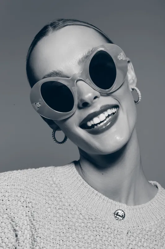 Young Caucasian woman with fair skin and slicked hair smiles wearing large sunglasses and hoop earrings in studio.