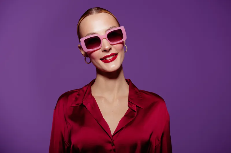 Stylish woman in red satin blouse and pink sunglasses smiles confidently against purple background. Fashion portrait with bright makeup and trendy accessories for modern style concept.