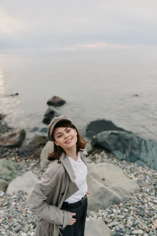 Smiling young woman in casual autumn outfit and hat stands on rocky beach near calm sea. Overcast sky creates soft natural light. Concept of leisure, travel, and outdoor lifestyle.