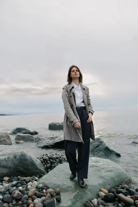 Young woman in a long coat stands on rocky shore with cloudy sky and calm sea in the background. Concept of fashion, autumn, and outdoor lifestyle with natural scenery.